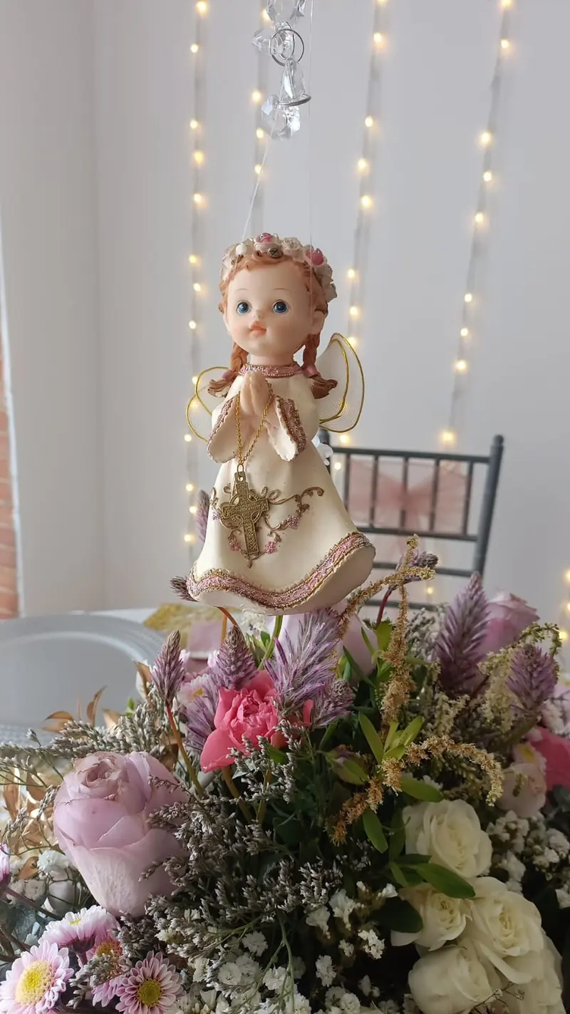 Centro de mesa de bautizo con flores frescas y una figura de ángel, frente a una cortina de luces