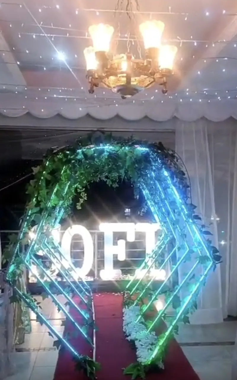 Arco de neón verde y azul sobre alfombra roja, con letras luminosas al fondo, en una fiesta temática